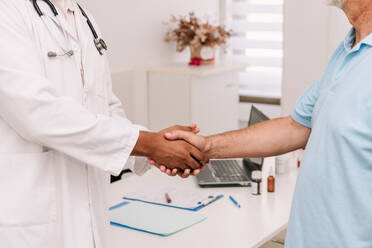 Side view of cropped unrecognizable senior male patient and African American doctor in medical uniform standing in hospital and shaking hands - ADSF47721