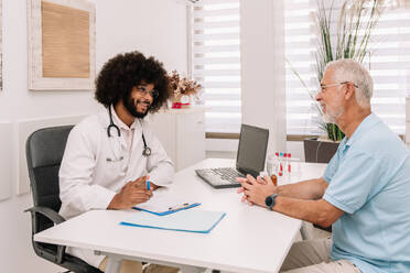 Cheerful multiracial doctor and patient sitting at table in hospital and talking to each other - ADSF47722