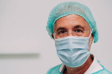 Positive mature male doctor in sterile uniform face mask looking at camera while standing in hospital - ADSF47726