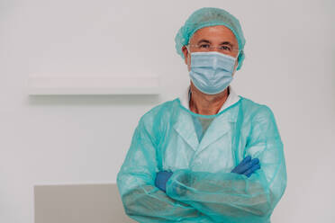 Positive mature male doctor in sterile uniform face mask and latex gloves looking at camera while standing in hospital with crossed arms - ADSF47727