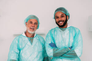 Cheerful multiracial bearded young and senior surgeons in sterile uniforms smiling and looking at camera after successful operation - ADSF47728