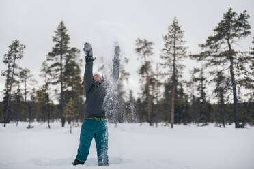 Full length of cheerful female in warm clothes throwing snow near trees in winter day in Lapland - ADSF47748