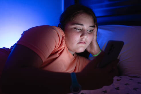 Side view of concentrated teenage girl looking at screen while lying on bed with a hand on his face in bedroom and watching scary movie on mobile phone in blue light - ADSF47756