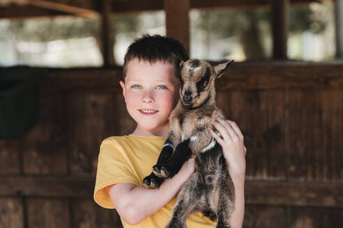 Portrait of smiling boy in casual clothes holding cute young American Pygmy while looking at camera in zoo - ADSF47760