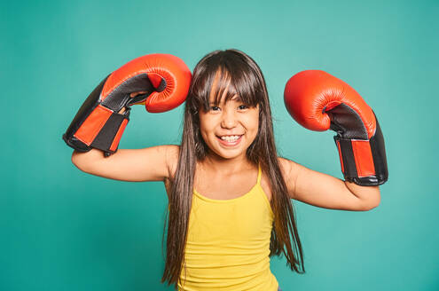 Portrait of smiling Asian girl with black hair and red boxing gloves looking at the camera while standing with her hands raised in a room against turquoise background - ADSF47766