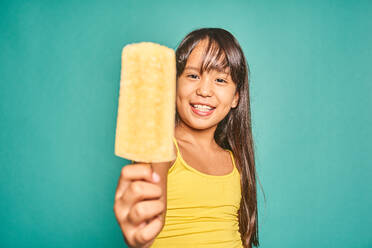 Cute Asian kid girl with bare shoulders looking at camera while standing and showing delicious ice cream with stick against turquoise background in light - ADSF47767