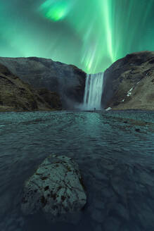 Picturesque scenery of waterfall flowing through mountains under green aurora borealis at night in Skogafoss Iceland - ADSF47796