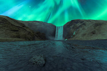 Picturesque scenery of waterfall flowing through mountains under green aurora borealis at night in Skogafoss Iceland - ADSF47797