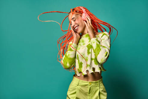 Young happy female in colorful outfit with orange Afro braids touching headphones while dancing with eyes closed to music against turquoise background - ADSF47802