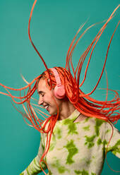 Young happy female in colorful outfit with orange Afro braids touching headphones while dancing to music against turquoise background - ADSF47803