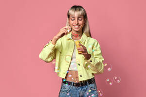 Young cheerful blond haired female with face piercings looking down in yellow jacket blowing soap bubbles from bottle while standing against pink background - ADSF47813