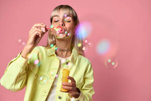Young blond haired female with eyes closed and face piercings in yellow jacket blowing soap bubbles from bottle while standing against pink background - ADSF47814