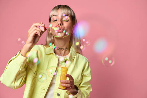 Young blond haired female with eyes closed and face piercings in yellow jacket blowing soap bubbles from bottle while standing against pink background - ADSF47814