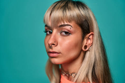 Portrait of young blonde woman with bangs and piercing on face wearing bright top looking at camera against turquoise background - ADSF47820