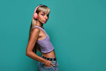 Side view of young woman looking at camera with blond hair wearing headphones while listening to song against turquoise background - ADSF47822
