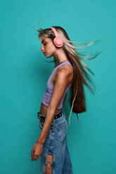 Side view of young woman with flying blond hair wearing headphones while listening to song against turquoise background - ADSF47823