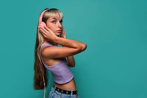 Side view of young woman looking away with blond hair wearing headphones while listening to song against turquoise background - ADSF47824