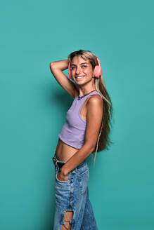 Side view of delighted young woman with blond hair wearing headphones while listening to song against turquoise background looking away - ADSF47825