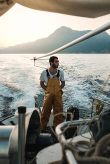 Nachdenklicher männlicher Fischer in Uniform in voller Länge, der beim Fischen in Soller in der Nähe der Baleareninsel Mallorca die Meereslandschaft bewundert - ADSF47832