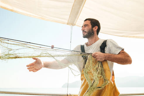 Fokussierter bärtiger männlicher Fischer in Uniform, der mit seinem Netz auf Fischfang geht, während er auf einem Schoner in Soller nahe der Baleareninsel Mallorca arbeitet - ADSF47845