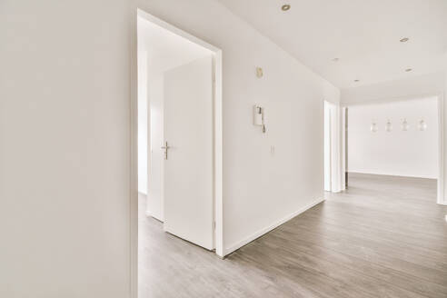 Narrow corridor with white walls and doors leading to spacious room with windows and parquet floor in modern apartment - ADSF47847