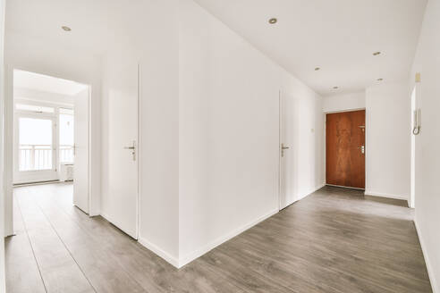 Narrow corridor with white walls and doors leading to spacious room with windows and parquet floor in modern apartment - ADSF47848