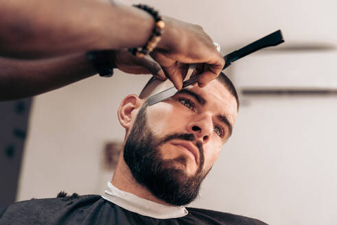 Crop anonymous professional barber shaving beard of brutal male client with straight edge razor in modern barbershop - ADSF47853