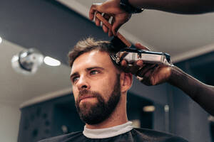 Crop anonymous barber using trimmer and brush while trimming hair to handsome bearded male client in barbershop against blurred background - ADSF47855