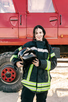 Junger positiver weiblicher Feuerwehrmann mit schwarzem Zopf, der einen Helm hält und eine Uniform trägt, die vor einem roten Feuerwehrauto steht - ADSF47856