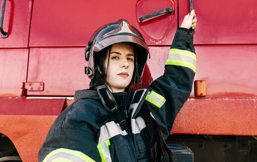 Junge furchtlose Feuerwehrfrau in Arbeitskleidung und mit Schutzhelm, die sich auf die Arbeit vorbereitet und in ein rotes Feuerwehrauto steigt - ADSF47857