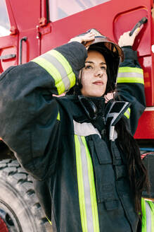 Junge furchtlose Feuerwehrfrau in Arbeitskleidung, die ihren Schutzhelm anpasst, während sie sich auf die Arbeit vorbereitet und in ein rotes Feuerwehrauto steigt - ADSF47859