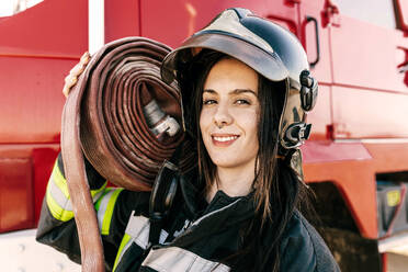 Tapferer weiblicher Feuerwehrmann mit Helm und Uniform, der einen schweren Schlauch auf der Schulter gegen ein rotes Feuerwehrauto trägt - ADSF47860