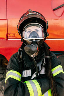 Female firefighter in professional uniform and protective respirator on head looking at camera standing with crossed arms near red truck - ADSF47861