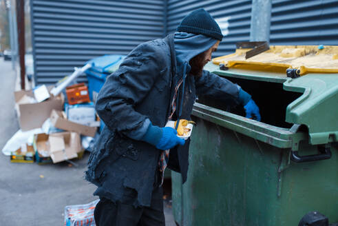 Männlicher bärtiger Bettler auf der Suche nach Essen in einem Mülleimer auf der Straße. Armut ist ein soziales Problem, Obdachlosigkeit und Einsamkeit, Alkoholismus und Trunksucht. Männlicher bärtiger Bettler auf der Suche nach Essen in einem Mülleimer - INGF12138