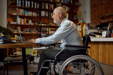 Adult disabled man in wheelchair using laptop, top view, disability, bookshelf and university library interior on background. Handicapped older male person, paralyzed people get knowledge. Adult disabled man using laptop, top view - INGF12165