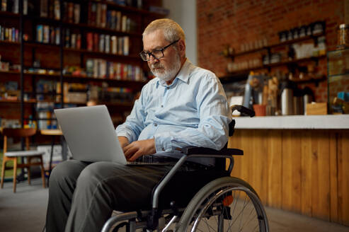 Adult disabled man in wheelchair using laptop, disability, bookshelf and university library interior on background. Handicapped older male person, paralyzed people get knowledge. Adult disabled man in wheelchair using laptop - INGF12166