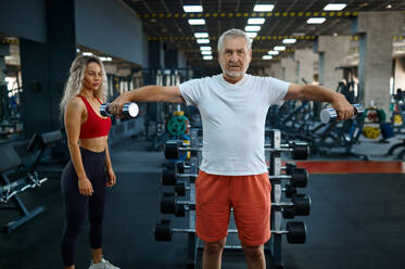 Old man doing exercise with dumbbells, female personal trainer, gym interior on background. Sportive grandpa with woman instructor, training in sport center. Old man, exercise with dumbbells, female trainer - INGF12167