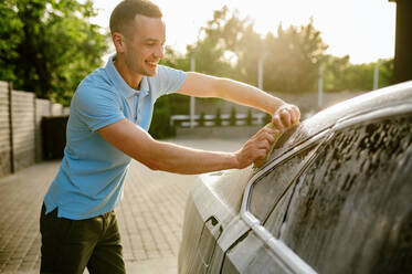 Man using sponge with foam, hand car wash station. Car-wash industry or business. Male person cleans his vehicle from dirt outdoors. Man using sponge with foam, hand car wash - INGF12171