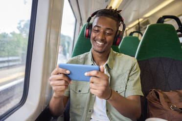 Young Man Commuting To Work Sitting On Train Streaming And Watching Film Or Show On Mobile Phone - INGF12185