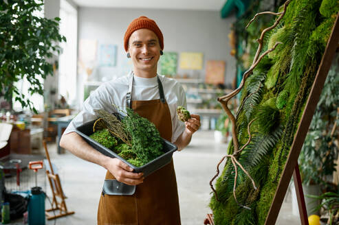 Portrait of young hipster florist smiling to camera standing at creative floral composition. Happy man decorator making decoration for home interior design. Portrait of young hipster florist standing at creative floral composition - INGF12186