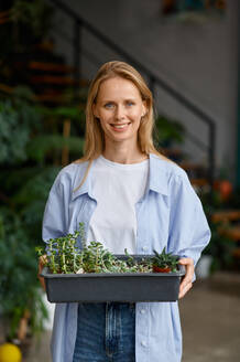 Smiling female florist worker enjoying her job. Portrait of smiling woman walking with plants in plastic box. Floristic workshop concept. Smiling female florist worker enjoying her job - INGF12192