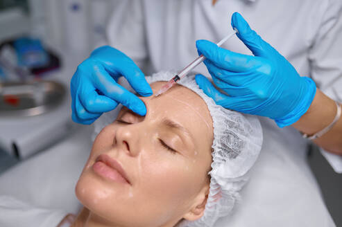 Anti-aging treatment and filler injection into eyebrow. Side view cosmetologist putting puncture of patient skin. Anti-aging treatment and filler injection between eyebrow concept - INGF12194