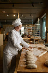 Young happy smiling woman baker in uniform forming bread loaves from raw dough at professional bakery kitchen. Woman baker forming bread loaves from raw dough at professional kitchen - INGF12203