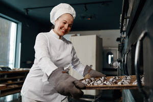 Happy smiling baker wearing white toque and jacket holding tray with cooked oatmeal cookies. Positive satisfied female chef taking hot fresh pastries out of oven. Happy smiling baker holding tray with cooked oatmeal cookies - INGF12205