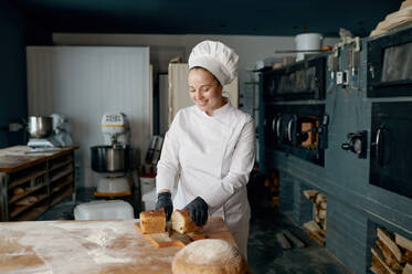 Young smiling woman baker chef holding knife and cutting fresh bread on wooden table. Healthy eating and traditional bakery concept. Baker chef holding knife and cutting fresh bread on wooden table - INGF12209