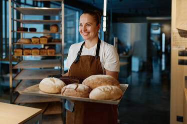 Pretty woman baker holding variety of baked bread on tray standing over bakery house background. Pretty woman baker holding variety of baked bread on tray - INGF12211