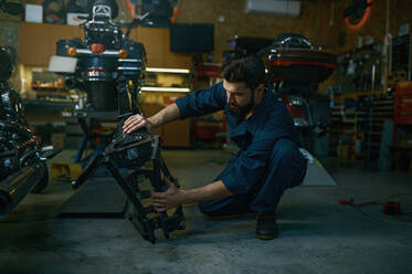 Young hipster man mechanic repairing motorcycle in workshop working with frame. Repairman checking and diagnosing parts on motorbike. Young hipster man mechanic repairs motorcycle in workshop working with frame - INGF12217