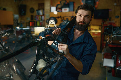 Portrait of young bearded repairman in coveralls carrying spare part of motorcycle frame on shoulder and looking at camera. Motorcycle garage repair service concept. Portrait of young repairman in coveralls carrying spare part of motorcycle - INGF12218