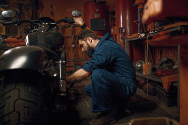 Mechanic working with motorcycle engine in workshop. Technician in blue workwear sitting nearby vehicle. Mechanic working with motorcycle engine in workshop while sitting nearby vehicle - INGF12221