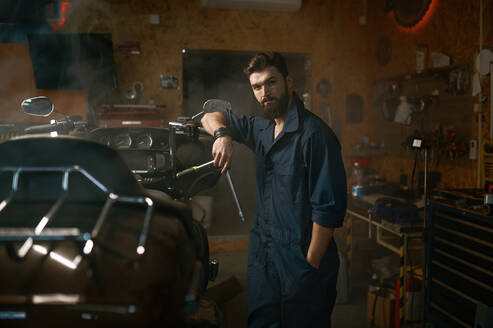 Handsome brutal repairman in work wear looking at camera portrait. Bearded young workman standing nearby motorcycle and holding wrench over smoky dark repair garage workplace. Handsome brutal repairman in work wear looking at camera portrait - INGF12223